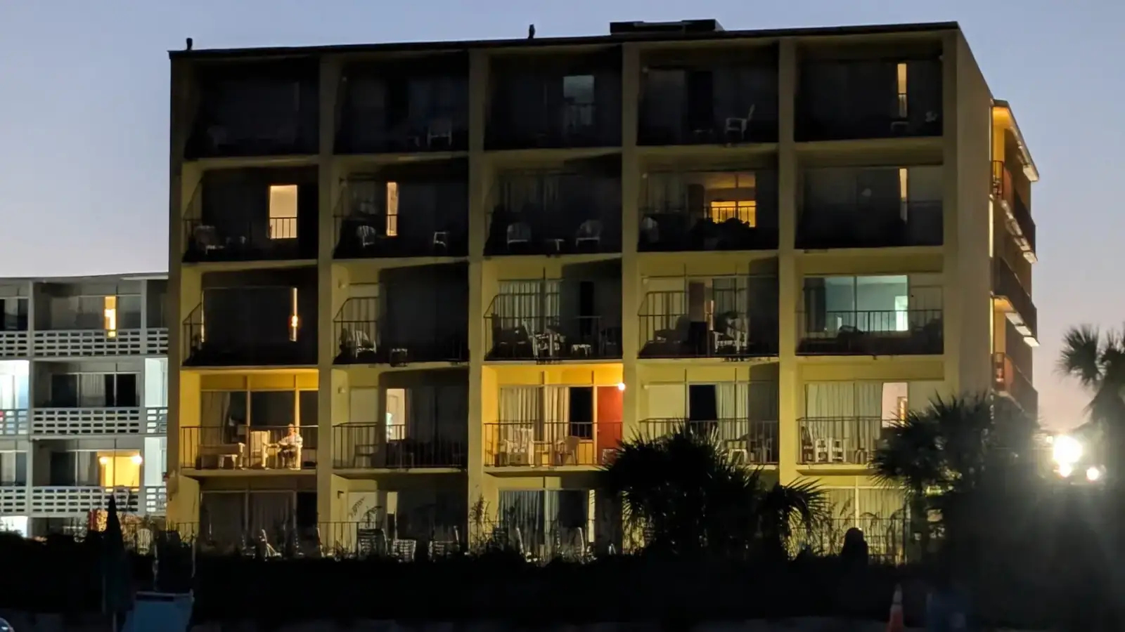 A low beachfront motel at dusk, one balcony lit gold with a man sitting in a chair